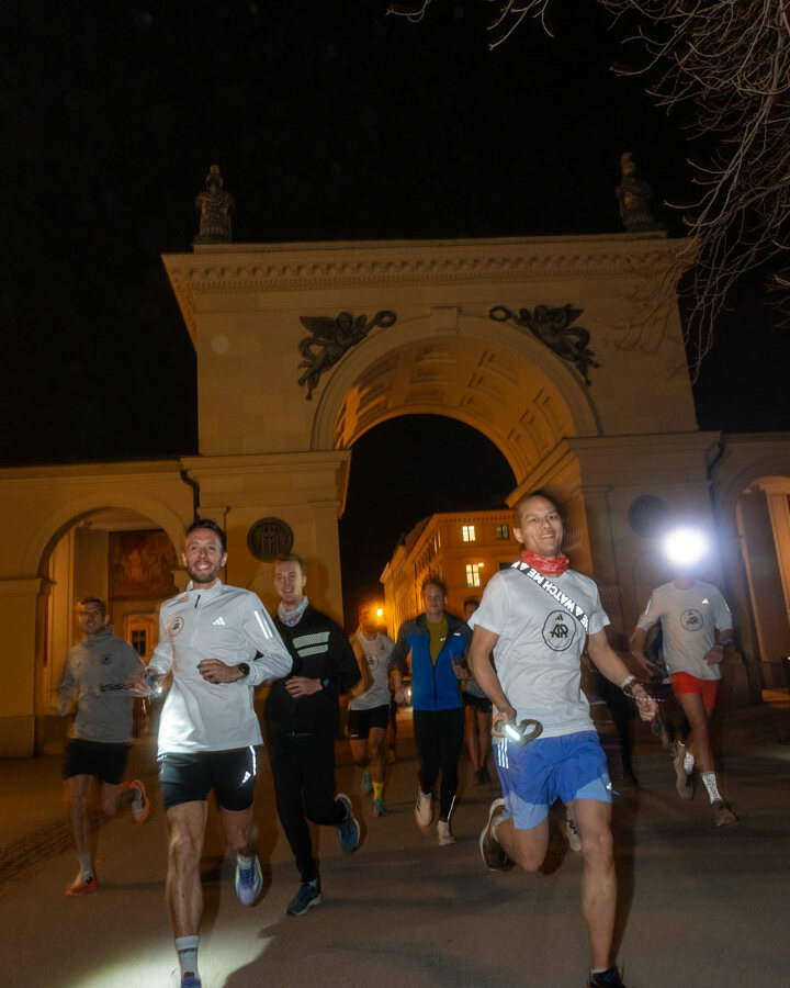 Eine Gruppe joggt nachts durch einen großen, beleuchteten Steintorbogen mit Statuen oben drauf und Bäumen und Gebäuden im Hintergrund – perfekte Bilder für eine Kampagne der Branding Agentur München oder des Social Media Management München.