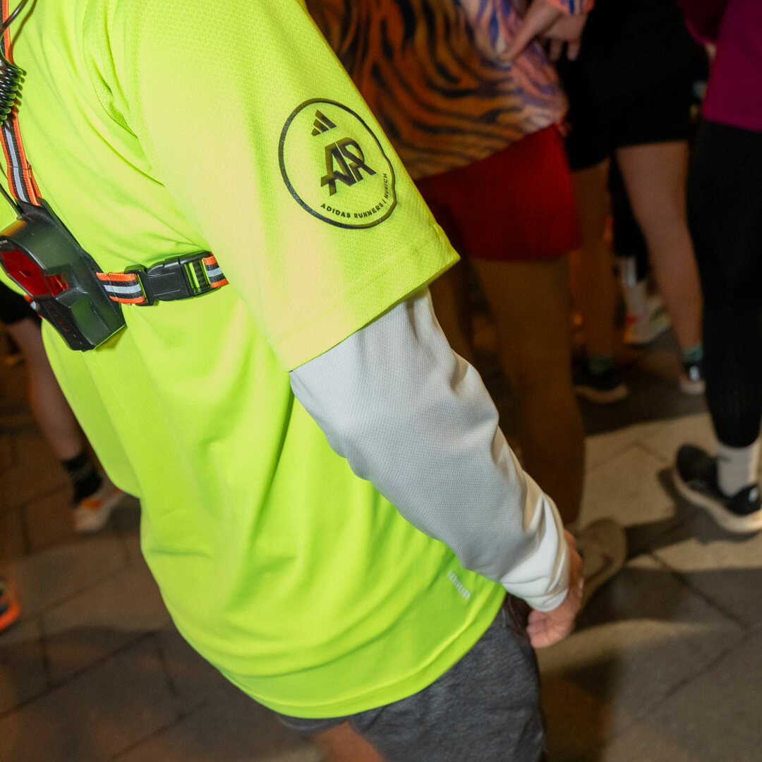Eine Person in leuchtend gelbem Shirt, grauen Shorts und rosa Socken steht inmitten einer Menschenmenge. Das Shirt ziert ein rundes Logo, die Person trägt einen Brustgurt mit einem kleinen Gerät – eine ideale Inspiration für die Produktion von Social Media Content.