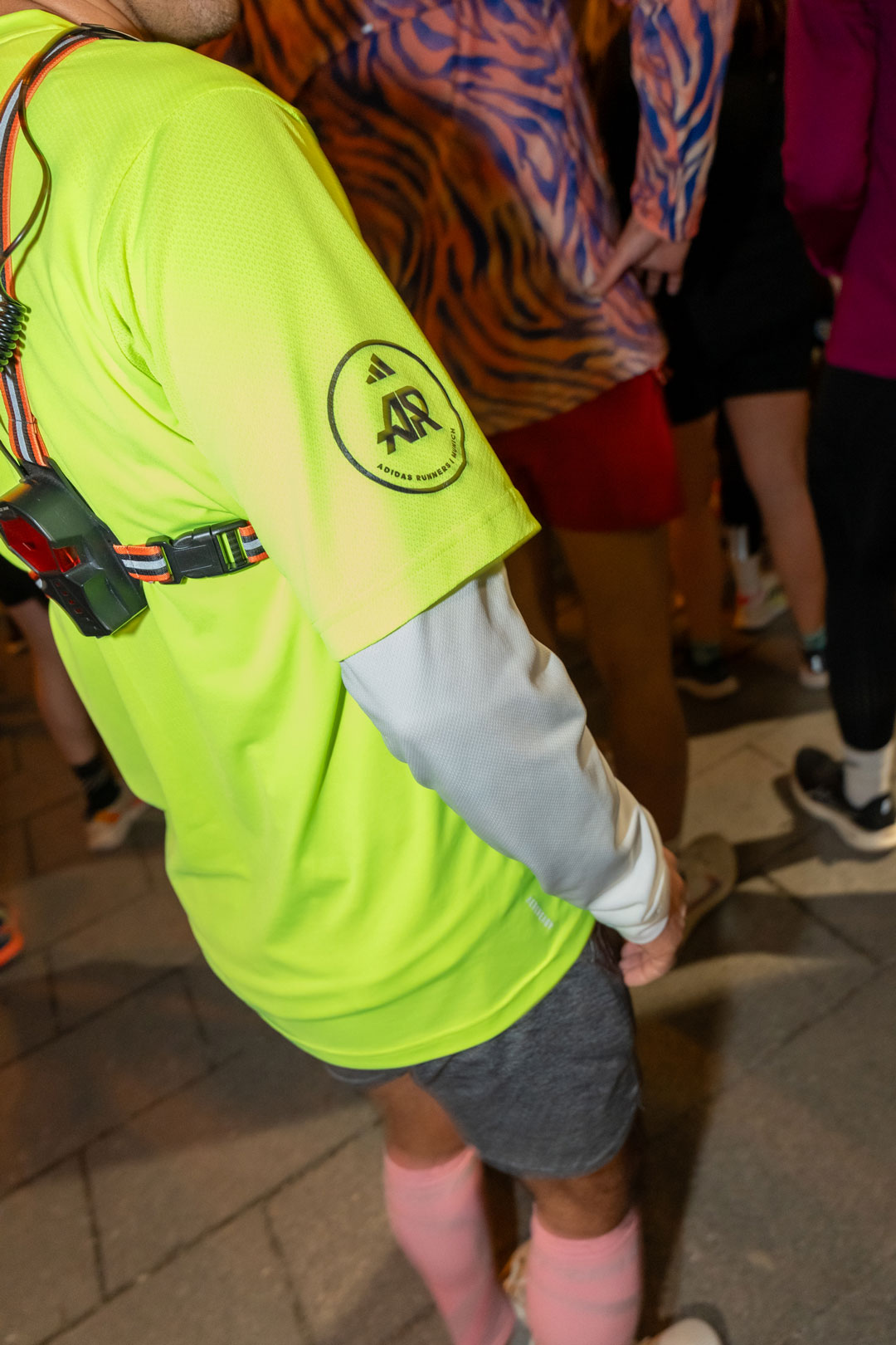 Eine Person in leuchtend gelbem Shirt, grauen Shorts und rosa Socken steht inmitten einer Menschenmenge. Das Shirt ziert ein rundes Logo, die Person trägt einen Brustgurt mit einem kleinen Gerät – eine ideale Inspiration für die Produktion von Social Media Content.