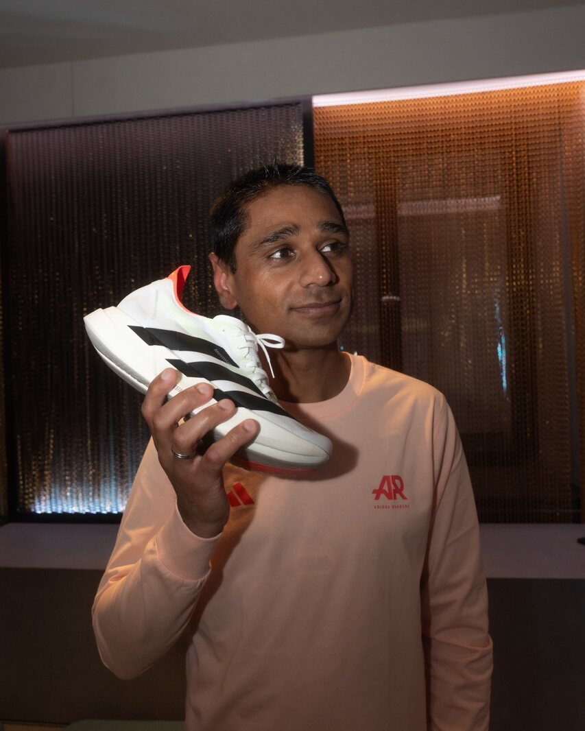 Ein Mann in einem hellrosa Sweatshirt hält einen weißen Sneaker mit schwarzen und roten Akzenten an sein Ohr, als würde er ihm zuhören, während er sich in Innenräumen bei sanfter Beleuchtung befindet – perfekt für die Produktion von Social Media-Inhalten oder innovative Kampagnen der Branding Agentur München.