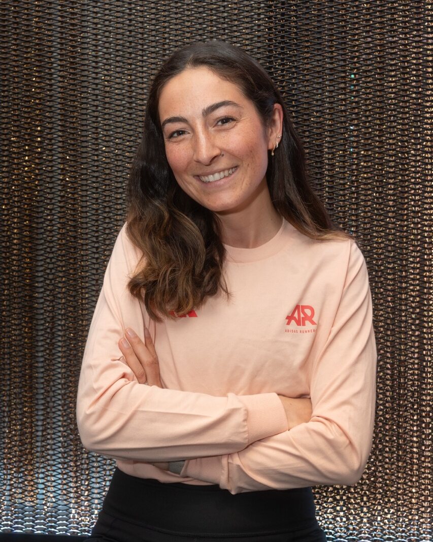 Eine Frau mit langen braunen Haaren, die ein hellrosa Langarmshirt und eine schwarze Hose trägt, sitzt lächelnd vor einem Perlenvorhang in die Kamera – perfekt für die Produktion von Social Media-Inhalten oder zur Präsentation Ihres Branding-Agentur-München-Stils.