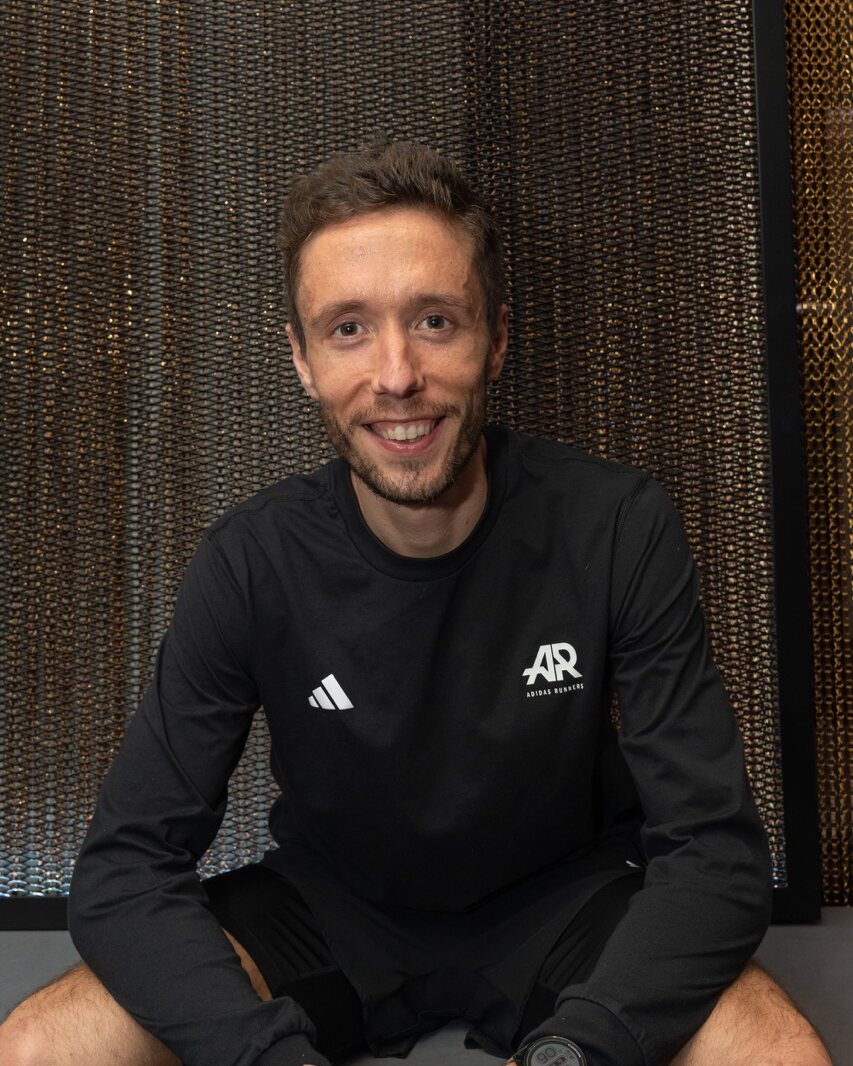 Ein Mann mit kurzen braunen Haaren und gestutztem Bart, der ein schwarzes Langarmshirt mit AR- und Adidas-Logos trägt, sitzt lächelnd vor einem strukturierten braunen Hintergrund – perfekt für Projekte von Content Creation München.
