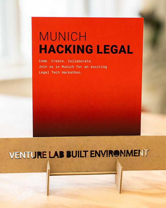 Auf einem rot-orangen Flyer auf einem Pappständer steht: MUNICH HACKING LEGAL. Coden. Kreieren. Zusammenarbeiten. Entdecken Sie die Zukunft von Legal Tech beim Eventvideo des Münchener Hackathons, veranstaltet von VENTURE LAB BUILT ENVIRONMENT.