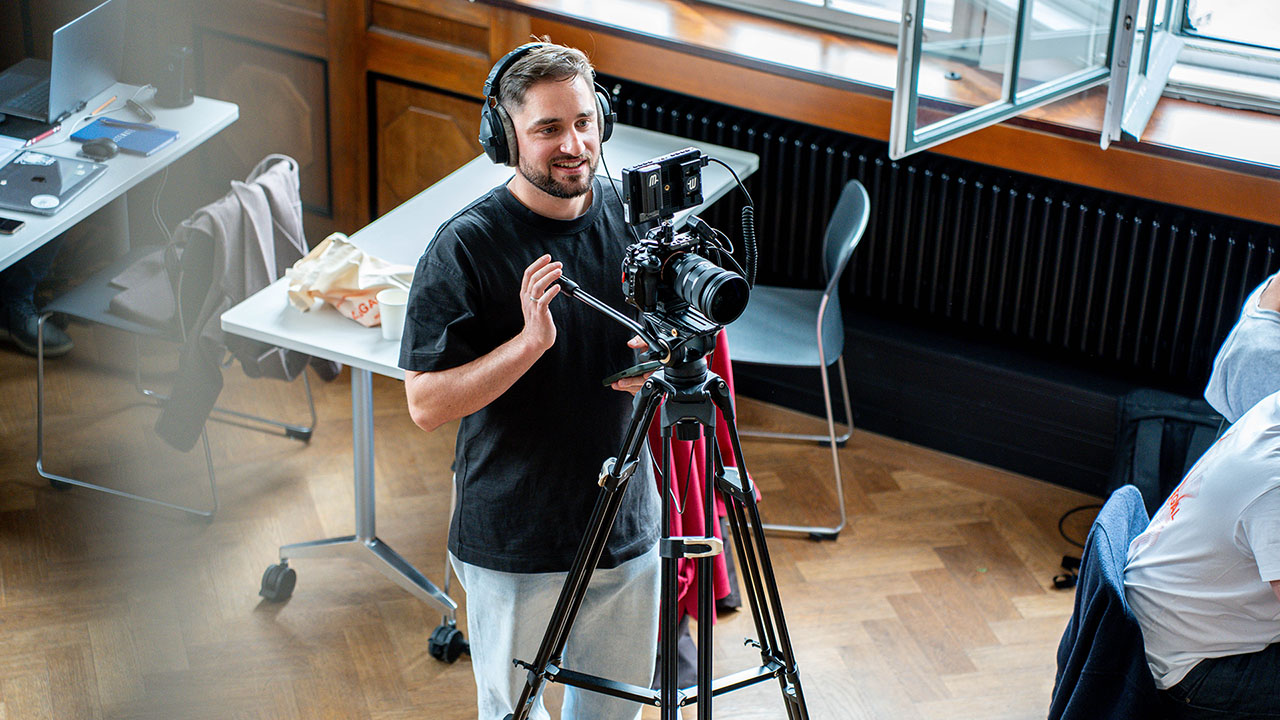 Thomas Widemann von WIDE Communication bedient eine Kamera auf einem Stativ in einem hellen Raum mit Holzboden, in der Nähe von Schreibtischen und einem offenen Fenster und erstellt Social-Media-Inhalte. Er scheint ein Videofilmer zu sein, der aufnimmt oder mit der Kamera spricht.